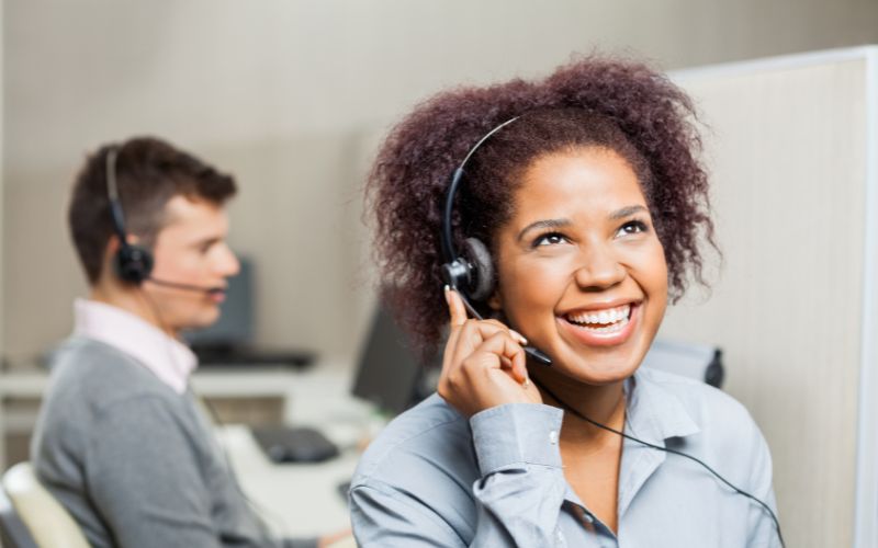 A personal cruise consultant with a radiant smile and headset engages in a friendly conversation, with a male colleague working in the background within a bustling cruise line call center environment.