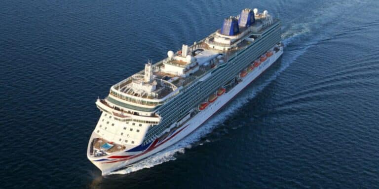 An aerial view of the P&O Britannia cruise ship sailing through the open sea. The ship's top deck features pools, sunbathing areas, and various amenities, while the Union Jack livery is prominently displayed on the bow. The vessel cuts smoothly through the water, leaving a wake behind.
