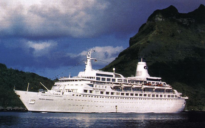 The Pacific Princess cruise ship, a large white vessel, is seen in clear waters with a mountainous backdrop. The ship's multiple decks and rows of windows shine under a cloudy sky, conveying a sense of adventure and luxury travel.