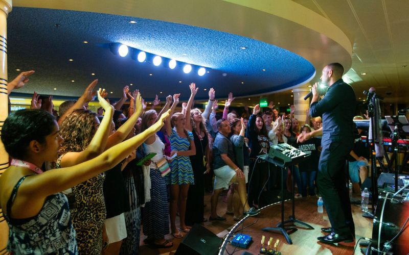 The image shows a lively scene of a trivia game taking place on a Carnival cruise ship. A host stands on stage, engaging with an enthusiastic crowd, many of whom have their hands raised. The atmosphere is energetic and fun, with the game taking place under bright indoor lighting.