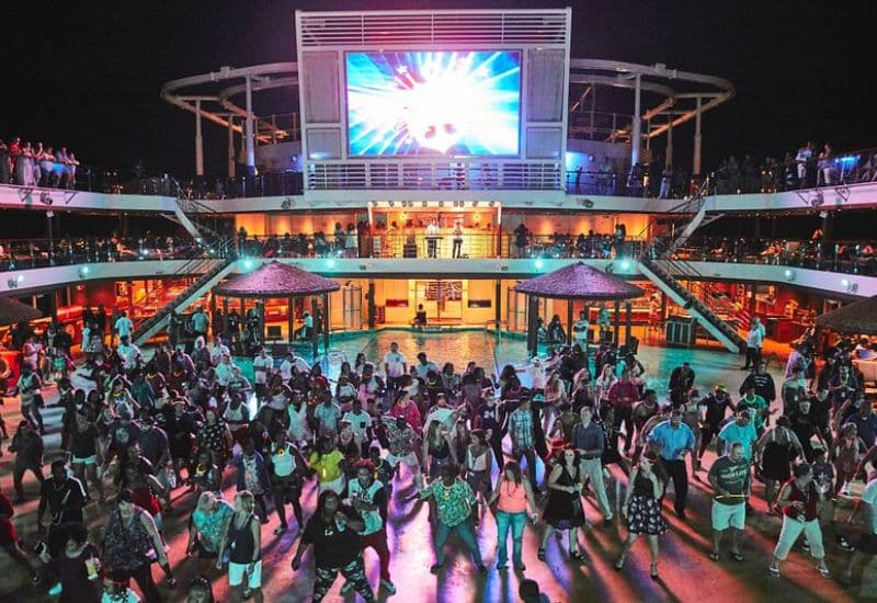 A vibrant dance party on a cruise ship deck at night, with a crowd of people dancing near the pool and a large screen illuminating the scene.