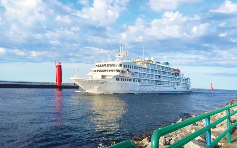 The Pearl Seas Cruises ship is anchored near a harbor, with a bright red lighthouse visible on the pier, against a backdrop of a serene sea and a partly cloudy sky, accented by the rocky shoreline in the foreground.