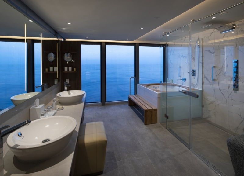 Luxurious Penthouse Suite bathroom on Celebrity Flora, with dual vessel sinks, a spacious glass-enclosed shower, and a Jacuzzi tub overlooking the ocean through floor-to-ceiling windows, combining modern design with breathtaking views.