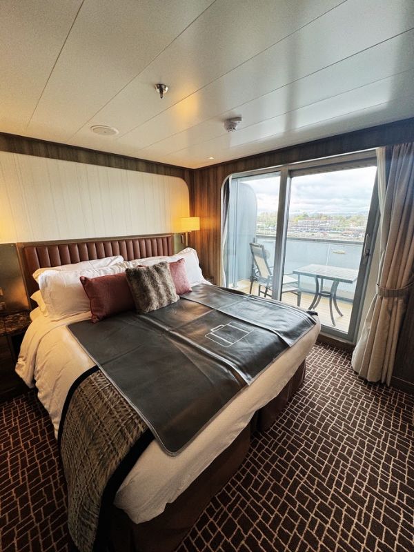 The bedroom area of the Penthouse Suite aboard the Seabourn Venture. The room is designed with a luxurious and comfortable king-sized bed, adorned with high-quality linens, a sleek grey throw, and plush decorative pillows. The bed is situated to allow guests to enjoy the stunning port views through the floor-to-ceiling sliding glass doors, which open onto a private verandah. The warm lighting, soft color palette, and elegant wood paneling create a serene and inviting atmosphere, perfect for relaxation after a day of exploration.