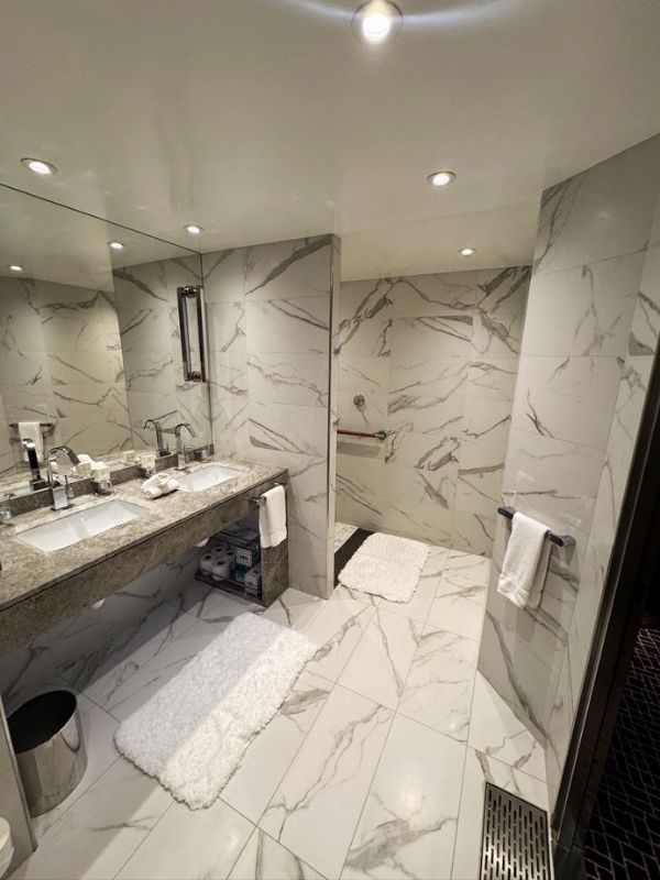 The luxurious bathroom in the Penthouse Suite on the Seabourn Venture. The bathroom is elegantly appointed with full marble walls and flooring, providing a sleek and sophisticated look. It features a large vanity with two sinks, a spacious mirror, and premium toiletries. Additionally, there is a generously sized walk-in shower and plush white towels, enhancing the spa-like feel of the space. The refined design and attention to detail in this bathroom ensure a lavish and comfortable experience for guests staying in the suite.