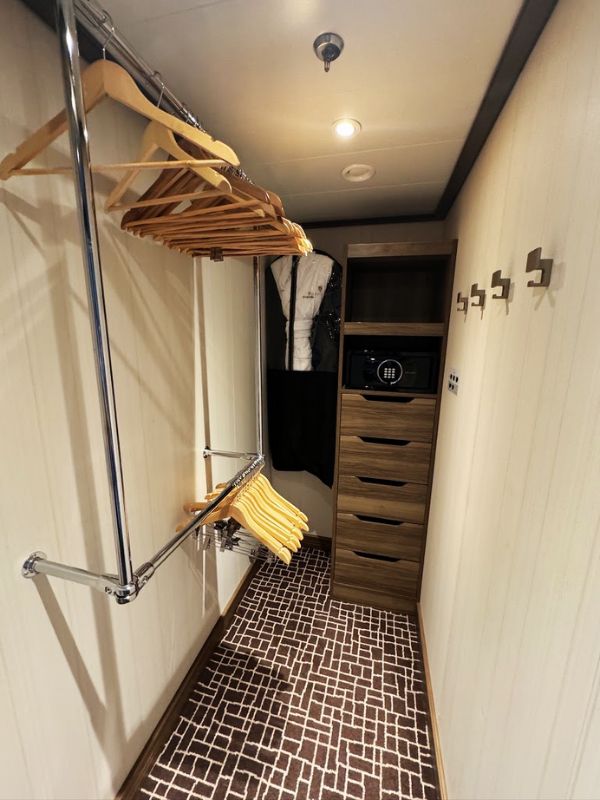 The spacious wardrobe area of the Penthouse Suite on the Seabourn Venture. It features a well-organized closet system with ample hanging space, accommodating a variety of clothing and accessories. Several wooden hangers are provided for guest use, ensuring garments can be stored neatly. Below the hanging area, a built-in dresser with multiple drawers offers additional storage options. This setup is practical yet elegant, designed to meet the needs of guests during their stay, providing convenience and a touch of luxury.