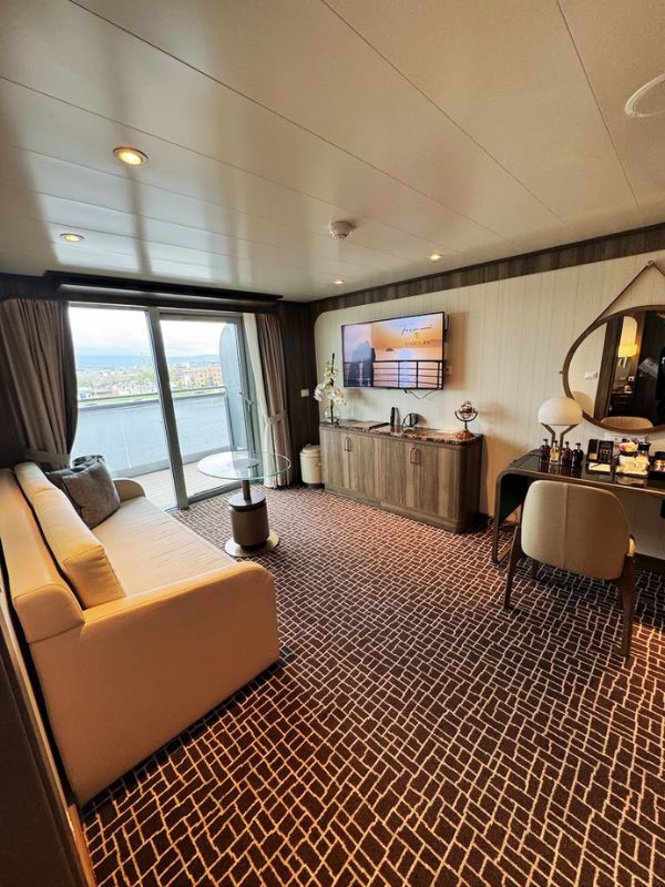 A view of the living area within the Penthouse Suite on the Seabourn Venture. The space is elegantly designed, featuring a large, plush sofa that faces a flat-screen TV mounted on a stylish decorative panel. Adjacent to the TV area is a small, round glass table and a modern chair, perfect for enjoying a drink or a quiet moment of reflection. The room is flooded with natural light from the large windows that offer stunning views of the port, enhancing the airy and spacious feel of the suite. This setting is not only luxurious but also incredibly comfortable, ensuring that guests feel at home while cruising.