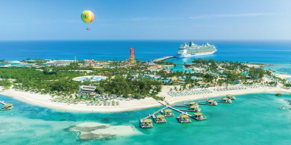 Aerial view of Perfect Day at CocoCay Island, showcasing the lush landscape, vibrant aqua park attractions, and a tethered yellow balloon, with a Royal Caribbean cruise ship anchored nearby.