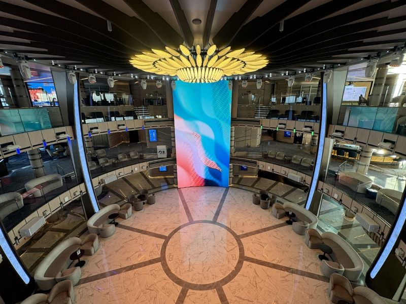 The image shows the Piazza on Princess Cruises' Sun Princess. This central atrium area features a stunning, large digital display that stretches from floor to ceiling, creating a focal point with vibrant, changing visuals. Surrounding the Piazza are several tiers of seating and viewing areas, arranged in a circular layout that gives the space an open, grand feel. The marble flooring and elegant furnishings, including curved seating and modern decor, enhance the luxurious atmosphere. Overhead, a striking chandelier adds to the opulence of the space, making the Piazza a key social and entertainment hub on the ship.