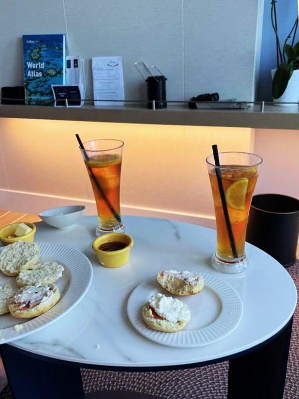 Two glasses of Pimm's and lemonade with lemon slices and straws, placed on a round white table with plates of scones topped with clotted cream and jam. The setting is a cozy lounge area on a cruise ship, with a world atlas and informational pamphlets on a shelf in the background.