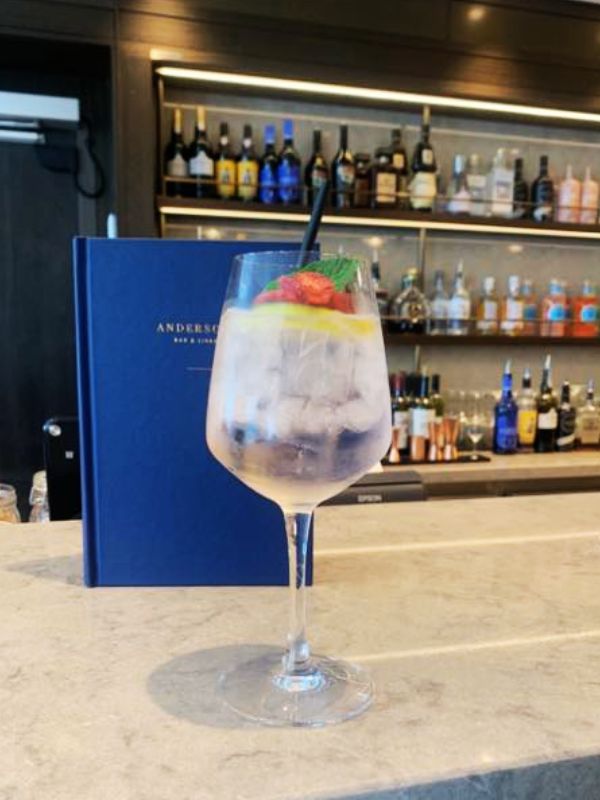 A Pink Gin Spritz on a P&O cruise ship, served in a large wine glass with ice, lemon slices, raspberries, and a mint sprig, complete with a straw. The drink is placed on a bar counter, with a blue menu and a well-stocked bar in the background, providing an elegant and inviting atmosphere for enjoying this refreshing cocktail.