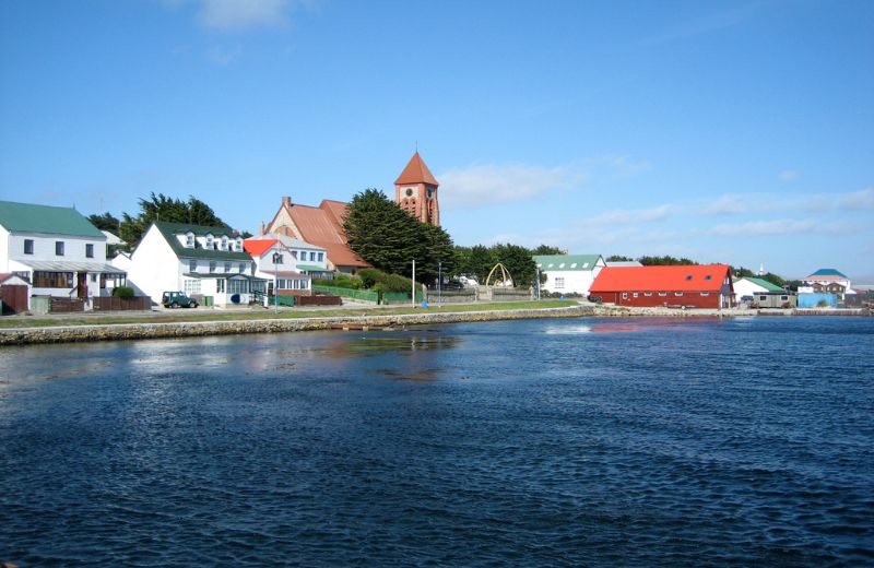 A serene view of Port Stanley, the capital of the Falkland Islands, featuring a tranquil waterfront scene. The town is depicted with charming, brightly colored buildings, including a prominent red-roofed church and traditional houses, against a clear blue sky. This small, picturesque settlement reflects a peaceful, remote island life, nestled along the edge of the calm waters, conveying a sense of isolation and quaint community living.