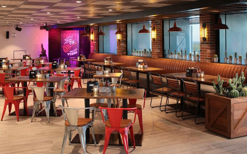 Interior of Portside BBQ restaurant featuring a cozy, modern dining area with a mix of red and silver chairs at wooden tables, a long brown leather booth, pendant lights, and exposed brick walls. A neon sign with the words 'Good Eats, Good Vibes' adds a vibrant touch to the ambiance."