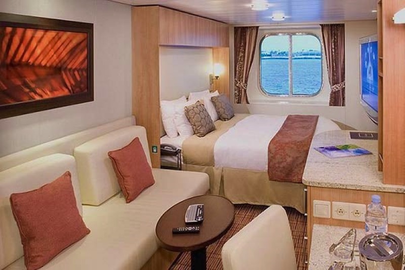 Cozy and inviting Premium Stateroom on the Celebrity Xpedition, featuring a comfortable bed, a sitting area with cream sofas, a round table, and an abstract painting, with a view of the ocean through a porthole.