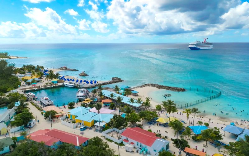 Elevated view of a Carnival cruise ship docked near Princess Cays, featuring the bustling tropical port with colorful cabanas, palm trees, clear turquoise waters, and guests enjoying the beachfront amenities.