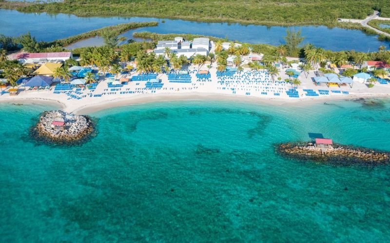 Aerial view of Princess Cays resort showing a bustling beach with rows of blue sun loungers under yellow umbrellas, a small pier, and a protective rocky breakwater creating a calm turquoise lagoon. The resort is a vivid contrast to the surrounding tranquil blue waters and green island vegetation.