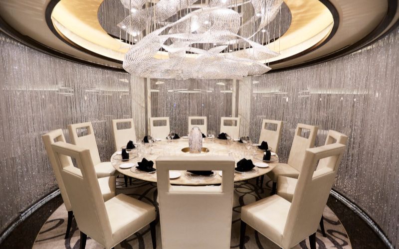 This image features an elegant dining space with a round table surrounded by high-backed, cream-colored chairs. The setting includes an opulent chandelier made of cascading crystals, creating a shimmering atmosphere. The backdrop features intricate, sparkling bead-like curtains, enhancing the luxurious ambiance.