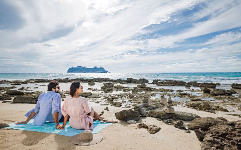 A couple sits on a sandy beach at Princess Cays, gazing out towards a Princess Cruises ship anchored in the distance. The rocky shoreline and tranquil sea create a serene backdrop, while the ship adds a sense of adventure to the peaceful scene.