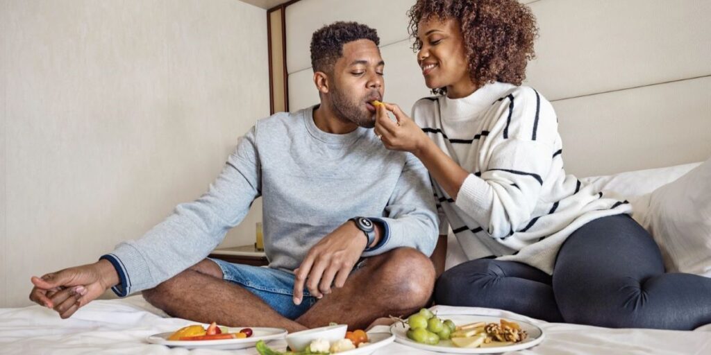 This image features a couple enjoying room service aboard a cruise, sitting on a neatly made bed. The woman playfully feeds the man while they share a spread of fresh fruit, vegetables, and other light snacks on white plates. The cozy, relaxed setting highlights the intimate and personalized dining experience offered in their cabin.