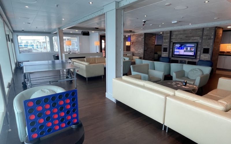 Teens club room on Princess Cruise Line, featuring comfortable beige sofas, a ping pong table, and a large Connect Four game. The room includes a TV area with a modern entertainment setup and offers a relaxed space for teenagers to socialize and enjoy various activities.