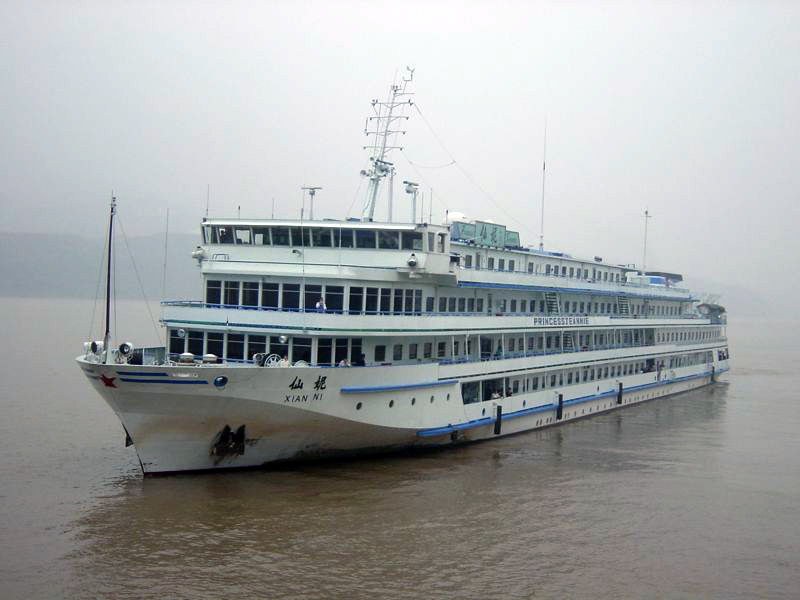 The 'Princess Jeannie' river cruise ship, a multi-deck vessel painted in white and blue, is captured navigating the murky waters of a wide river on an overcast day.