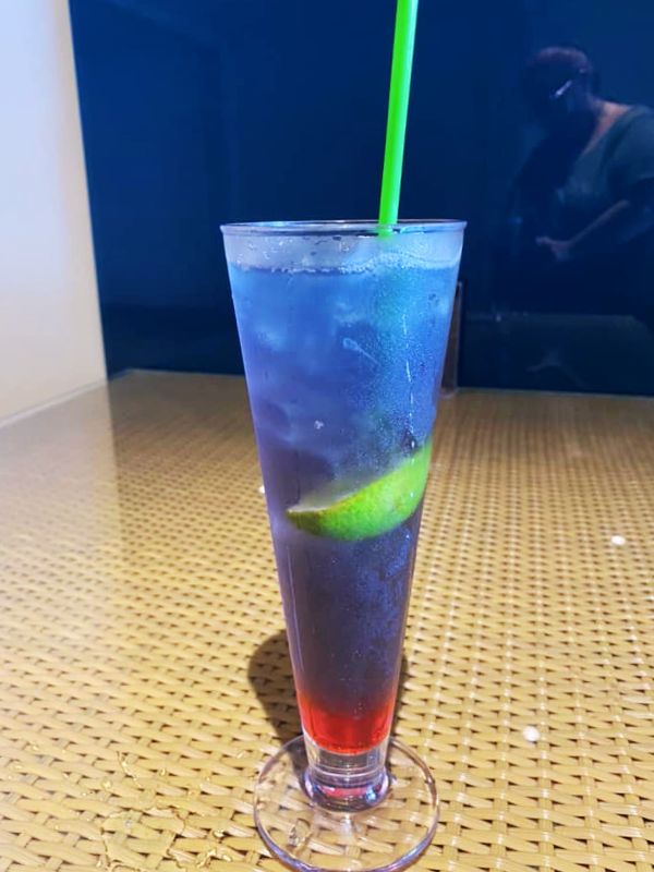 A Purple Rain cocktail on an MSC Cruises ship, presented in a tall glass with layers of vibrant purple and red hues, garnished with a lime wedge and served with a green straw. The drink is placed on a woven table, with a reflective background adding to the sleek and stylish presentation.
