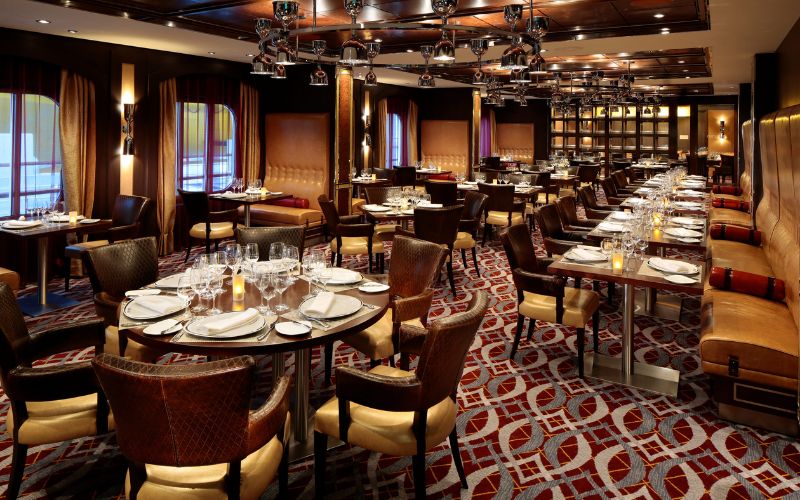 Elegant dining at Chops Grille on Royal Caribbean's Quantum of the Seas, featuring a sophisticated setup with polished wood tables, leather chairs, fine china, and mood lighting, inviting guests to a premium steakhouse experience at sea.