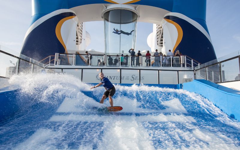 A guest enjoying the FlowRider surf simulator aboard Quantum of the Seas, with onlookers observing from behind a clear viewing panel and a person suspended in the skydiving simulator above, all set against a backdrop of the ship's distinctive blue and yellow funnel.