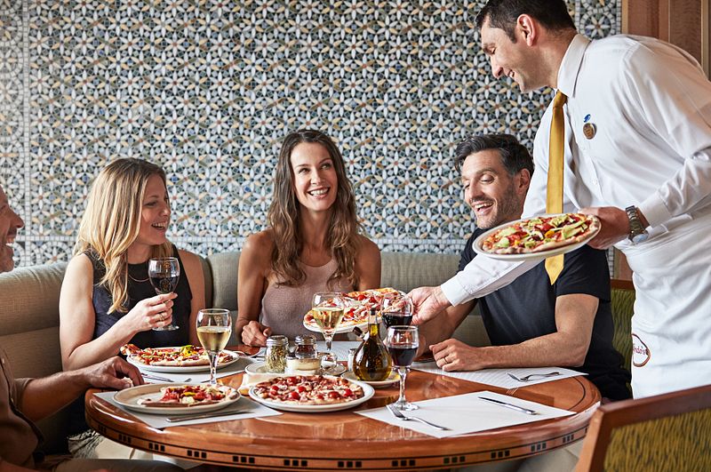 Guests enjoying a delightful dining experience at Alfredo's Pizzeria on Royal Princess, with a waiter serving a fresh, hot pizza. The atmosphere is casual yet elegant, as the diners engage in cheerful conversation, with wine glasses and a variety of pizzas on the table, accentuating the restaurant's cozy and inviting ambiance.