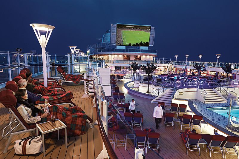 Guests cozily wrapped in plaid blankets on deck chairs enjoy 'Movies Under the Stars' on a Princess cruise ship. The evening atmosphere is enlivened by a large outdoor screen, with the ship's pool and open decks illuminated under the night sky.