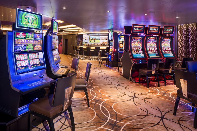 Inside the casino on Holland America Line's Rotterdam, showcasing a range of slot machines with bright screens, elegant chairs, and a sophisticated ambiance with stylish carpeting.