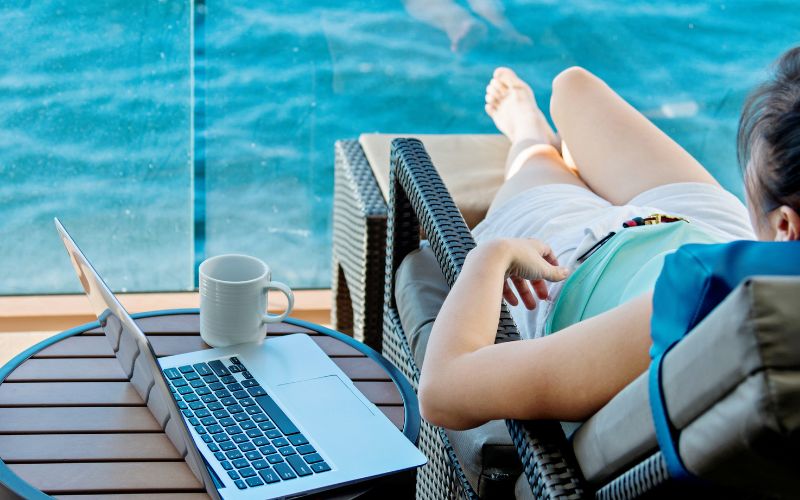 A lady relaxing on the balcony of a criuse next to a table with a drink and a laptop.