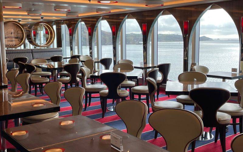 Elegant dining area inside a Holland America Line cruise ship with modern beige and black chairs, red carpet flooring, and large windows offering a panoramic view of the sea.