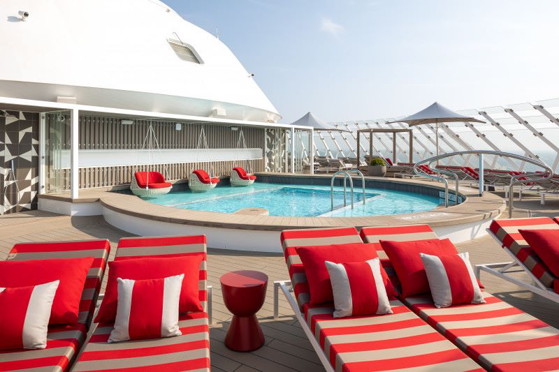 The exclusive Retreat Sundeck on Celebrity Beyond, featuring a serene pool area with luxurious red and white loungers, matching pillows, and side tables, all set against a backdrop of modern design elements and open skies.