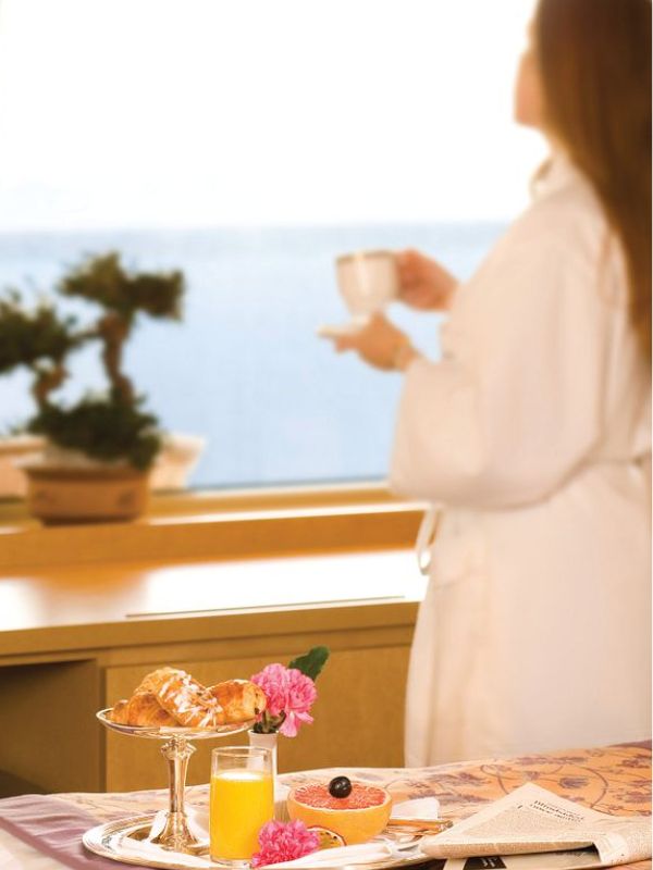 A serene room service setting on a Holland America Line cruise ship, with a guest in a white robe enjoying a cup of coffee by a window overlooking the sea, alongside a tray with a glass of orange juice, pastries, fresh fruit, and flowers.