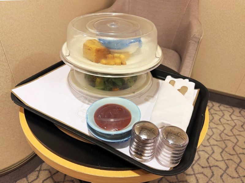 A room service tray on a Cunard cruise ship, featuring covered plates with food, a bowl of sauce, salt and pepper shakers, and neatly arranged napkins and cutlery on a small table.