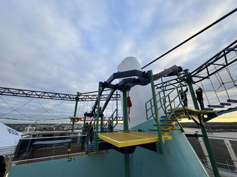 A close-up of the adventure ropes course on MSC Euribias top deck, featuring various challenging elements and safety harnesses against a cloudy sky, with a cityscape and river visible in the background.