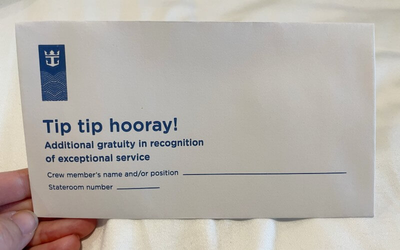 A hand holds a tip envelope from a cruise ship with the text "Tip tip hooray! Additional gratuity in recognition of exceptional service." The envelope includes fields for the crew member's name or position and the stateroom number.