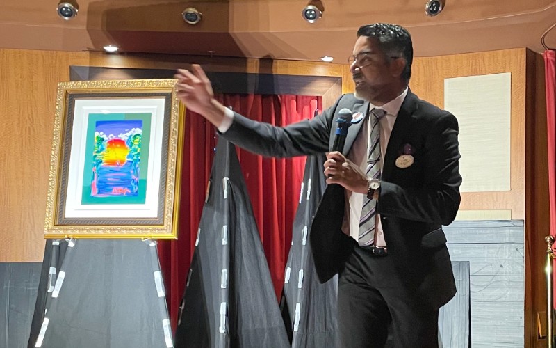 An art auctioneer on Royal Caribbean's cruise ship engaging with guests during an art auction, gesturing towards a colorful framed artwork, providing an entertaining and cultured experience at sea.