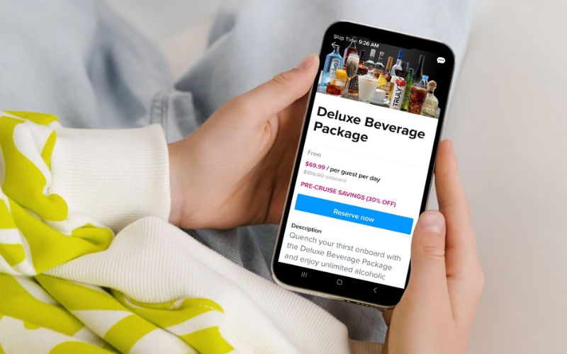 Close-up of a person's hand holding a smartphone displaying a cruise ship's Deluxe Beverage Package offer, with a variety of alcoholic drinks in the background on the screen, suggesting the convenience of mobile booking.