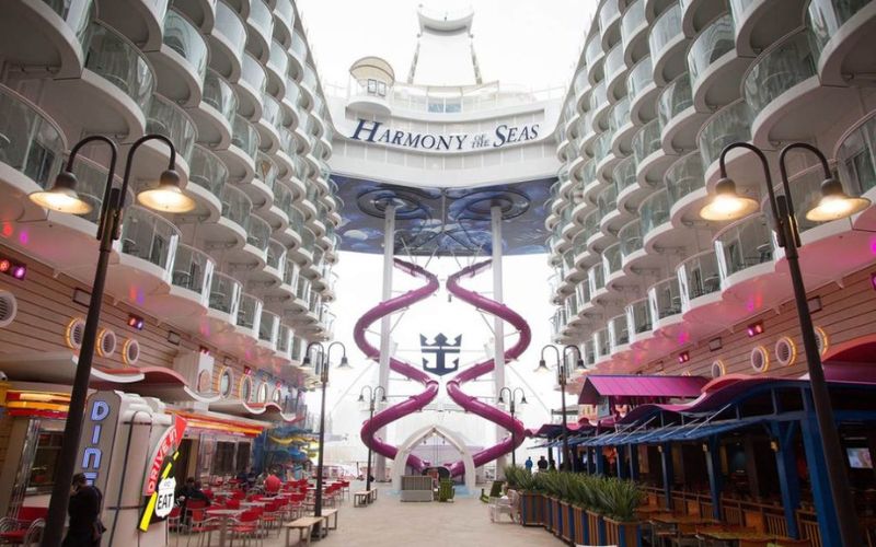 The Ultimate Abyss, the tallest slide at sea, descends dramatically on Royal Caribbean's Harmony of the Seas, set against the backdrop of the ship's unique architecture with colorful Boardwalk eateries and ambient lighting.