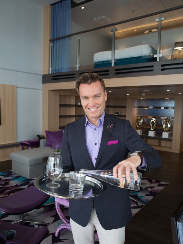 A smiling Royal Genie in a smart purple and navy uniform, serving water in the luxurious Royal Loft Suite on Harmony of the Seas, with the suite's elegant interior in the background.