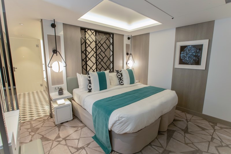 The Royal Suite on a Celebrity cruise ship, with a modern and inviting design featuring a comfortable bed with a white and teal color scheme, elegant wall lamps, a decorative lattice headboard, and an art piece that adds a touch of sophistication to the room.