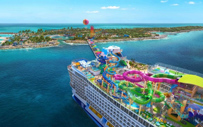 A vibrant aerial view of a Royal Caribbean cruise ship, featuring an elaborate top deck with colorful water slides and leisure areas, approaching a tropical island with clear blue waters and sandy beaches, under a bright blue sky.