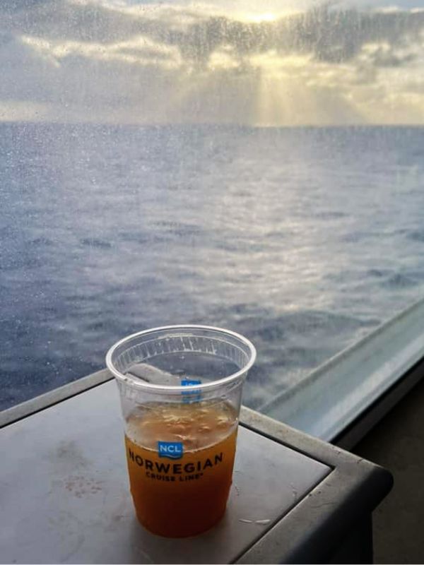 A Rum Swizzle cocktail in a clear plastic cup with the Norwegian Cruise Line logo, placed on a ledge overlooking the ocean. The background features the expansive sea and sky, creating a serene and relaxing ambiance typical of a cruise experience.