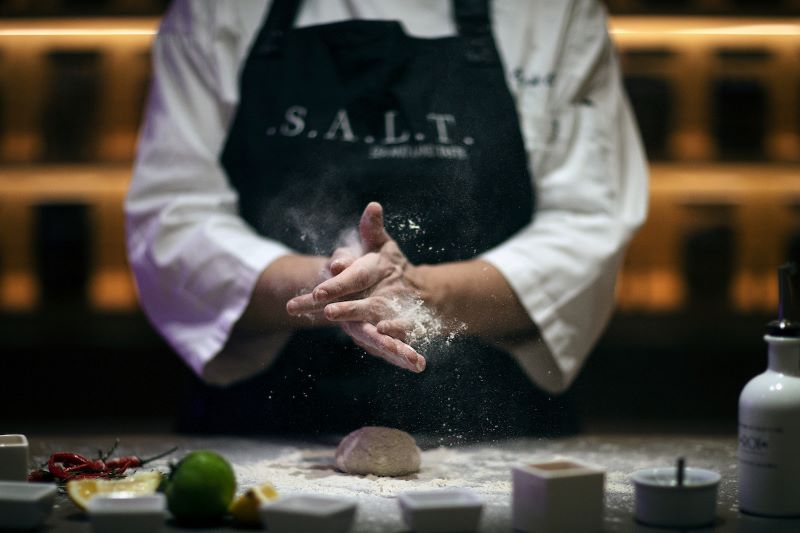 A chef at the S.A.L.T. Chef’s Table on a Silversea cruise ship, captured in a dynamic moment of culinary preparation, with flour being expertly sprinkled onto dough amidst a backdrop of ingredients and a professional kitchen setting.