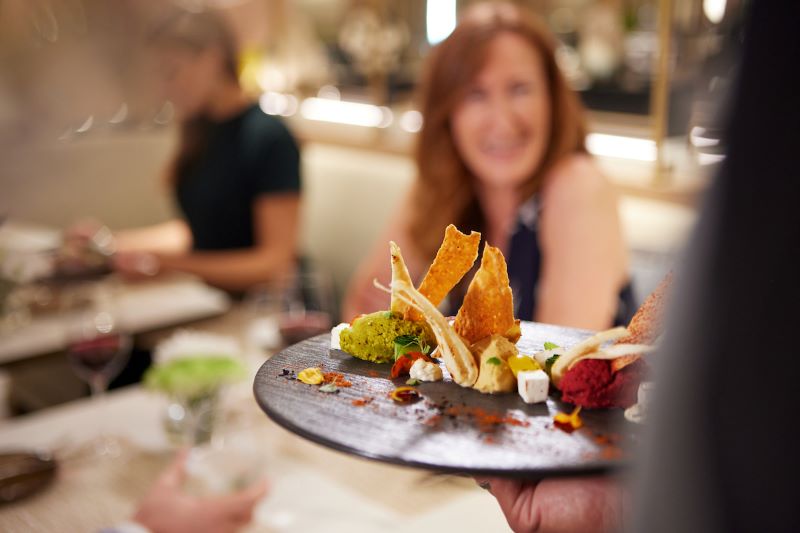 A gourmet dish is presented at the S.A.L.T. Kitchen on a Silversea cruise ship, with focus on the artfully arranged cuisine, while a smiling guest in the softly blurred background anticipates the dining experience.