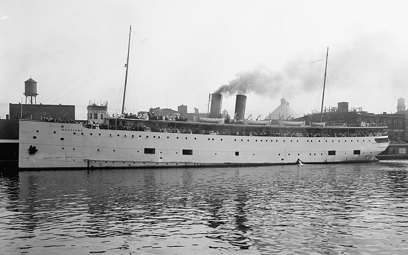 Historic image of the SS Eastland, a passenger ship, moored in a harbor with its deck crowded with passengers, smoke rising from one of its two funnels, reflecting the bustling maritime activity of the era.