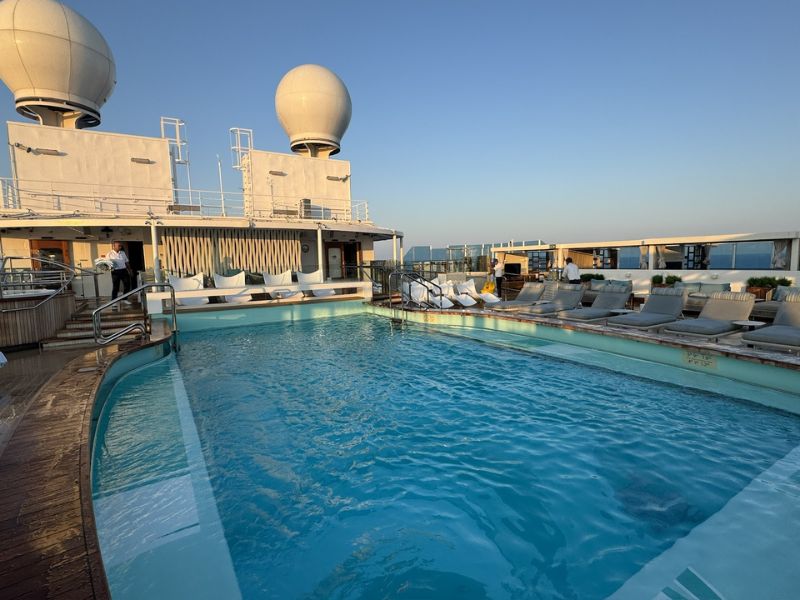 The image shows the Sanctuary Sun Deck on Princess Cruises' Sun Princess. This area features a tranquil swimming pool surrounded by lounge chairs, offering a relaxing space for guests to unwind. The deck includes cabana-style seating areas with white cushions, adding a touch of luxury to the environment. The pool area is framed by two large radar domes on the upper deck, and the clear sky suggests a serene and peaceful atmosphere. The overall design emphasizes comfort and exclusivity, making it an ideal spot for relaxation.