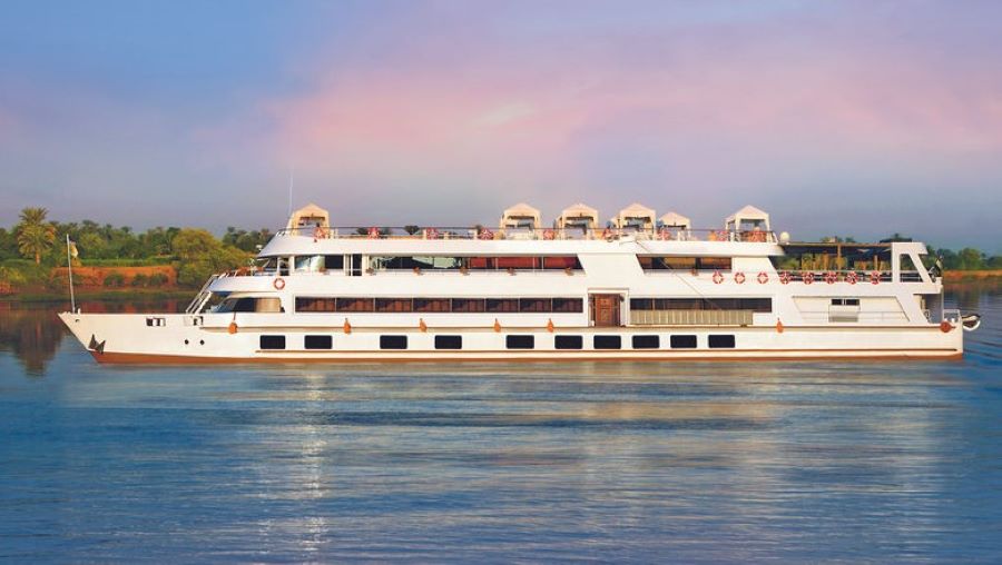 A Scenic luxury river cruise ship glides on calm waters during a pastel-hued sunset. The ship's white and gold trimmings gleam, and the top deck with lounge chairs is invitingly visible. Gentle ripples in the water and the silhouette of palm trees on the distant shore complete this idyllic river cruising moment.